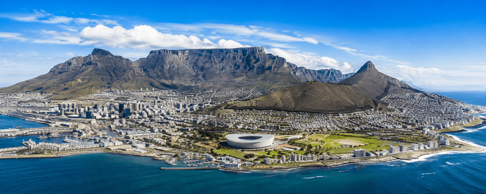 Panoramic daytime view of Cape Town with Table Mountain, the city skyline, coastline and Atlantic Ocean beneath a bright blue South African sky.